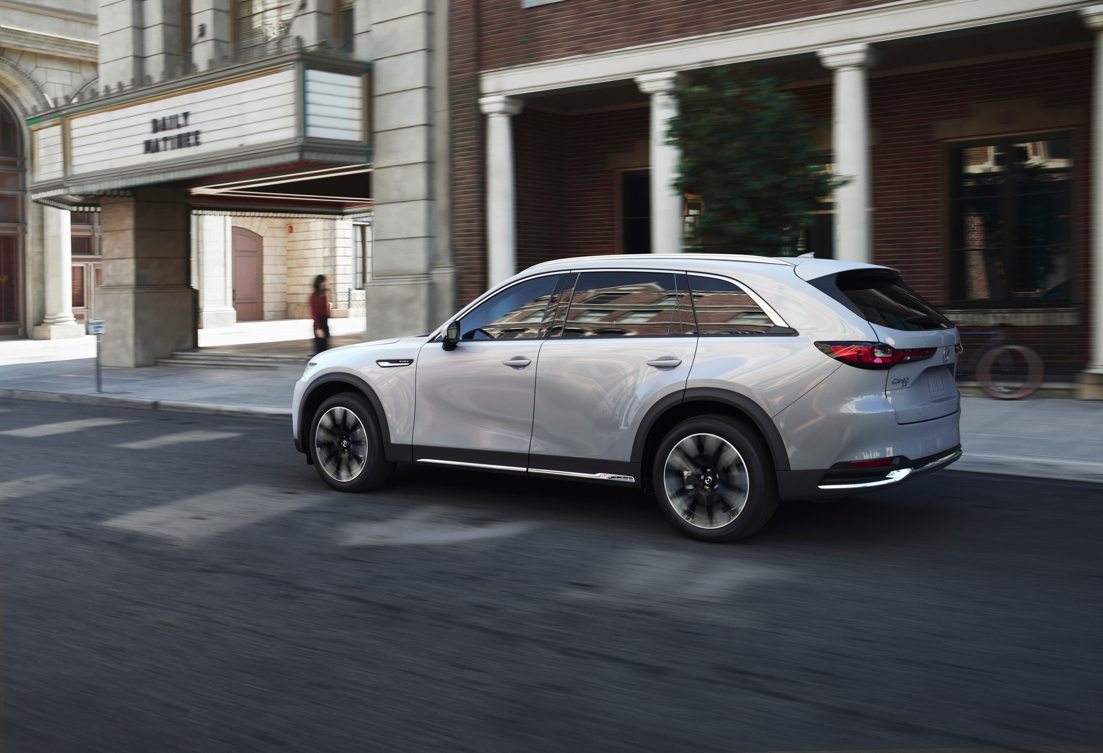 A white CX-90 plug-in hybrid moving along a city street, its PHEV badge visible on the front wing.
