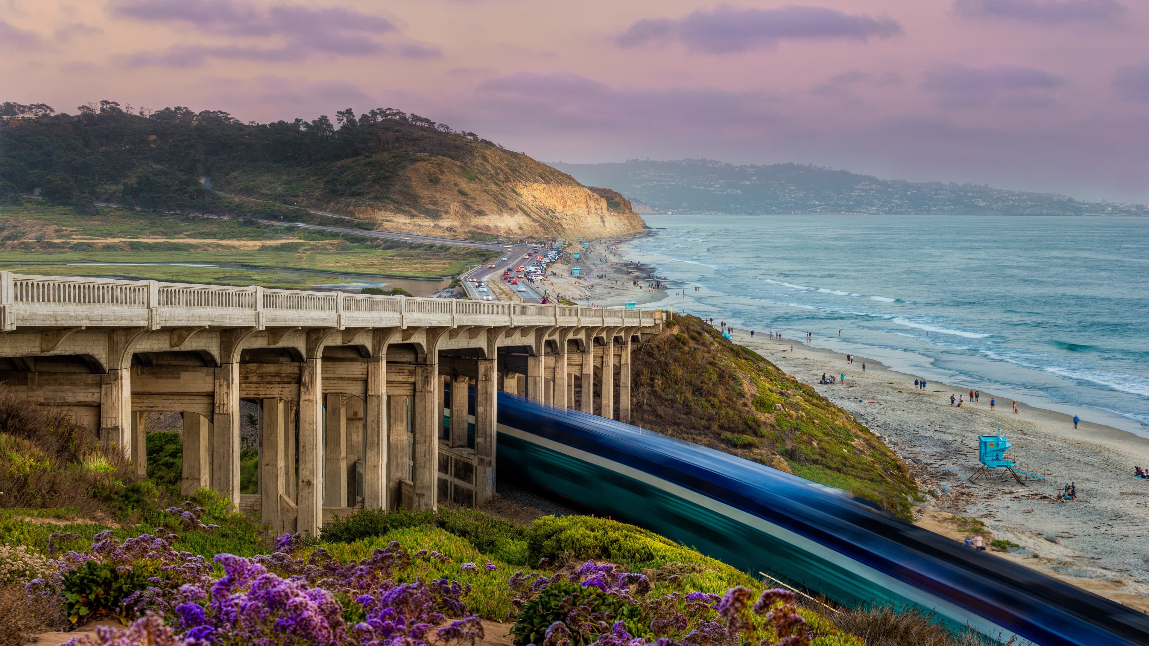 Cars driving south on Highway 101 past Torrey Pines State Beach, with ocean views and coastal train in background, an iconic San Diego route.