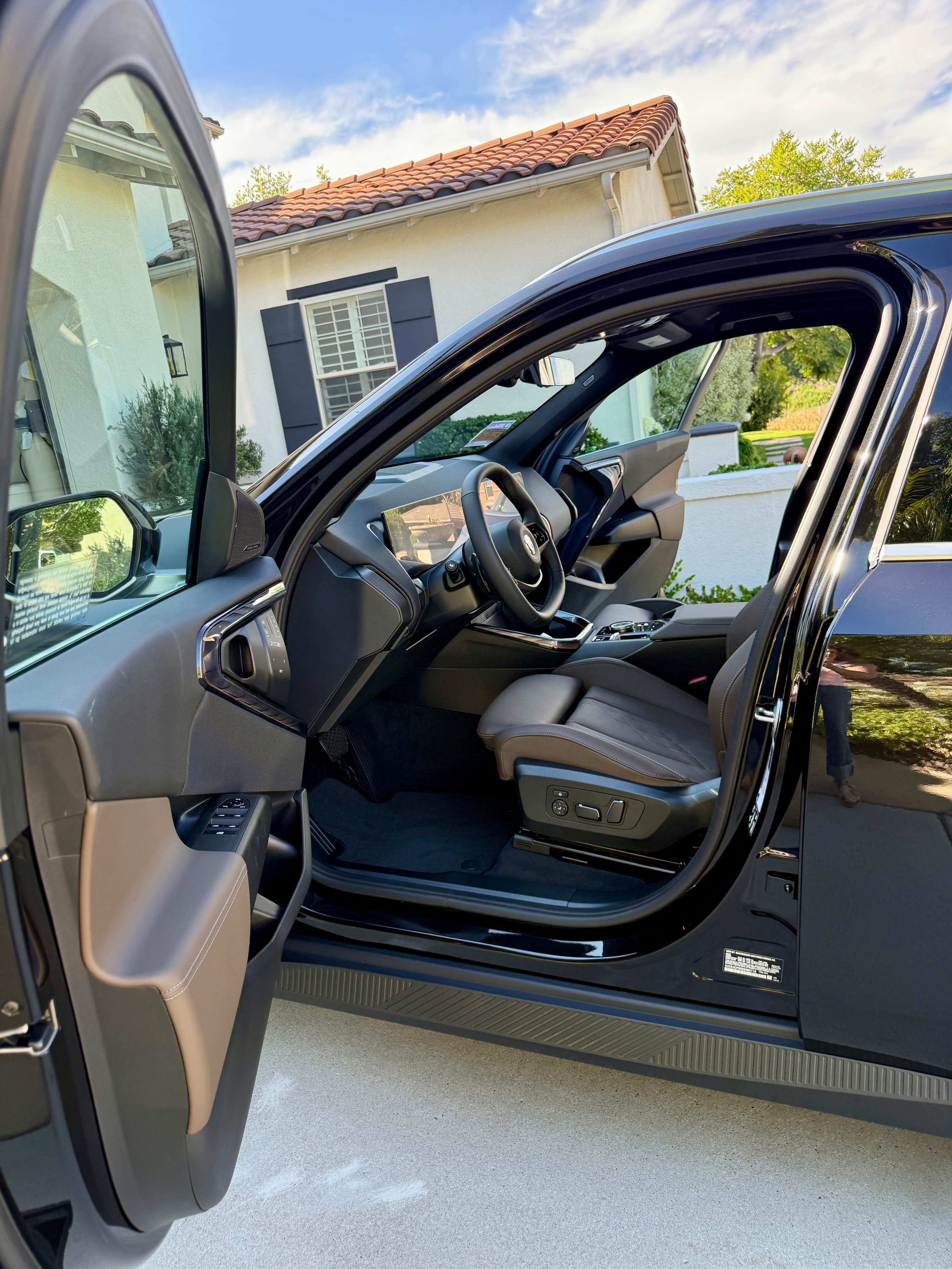 The open driver’s door of a black BMW on a residential driveway, showing the brown leather interior and the dashboard